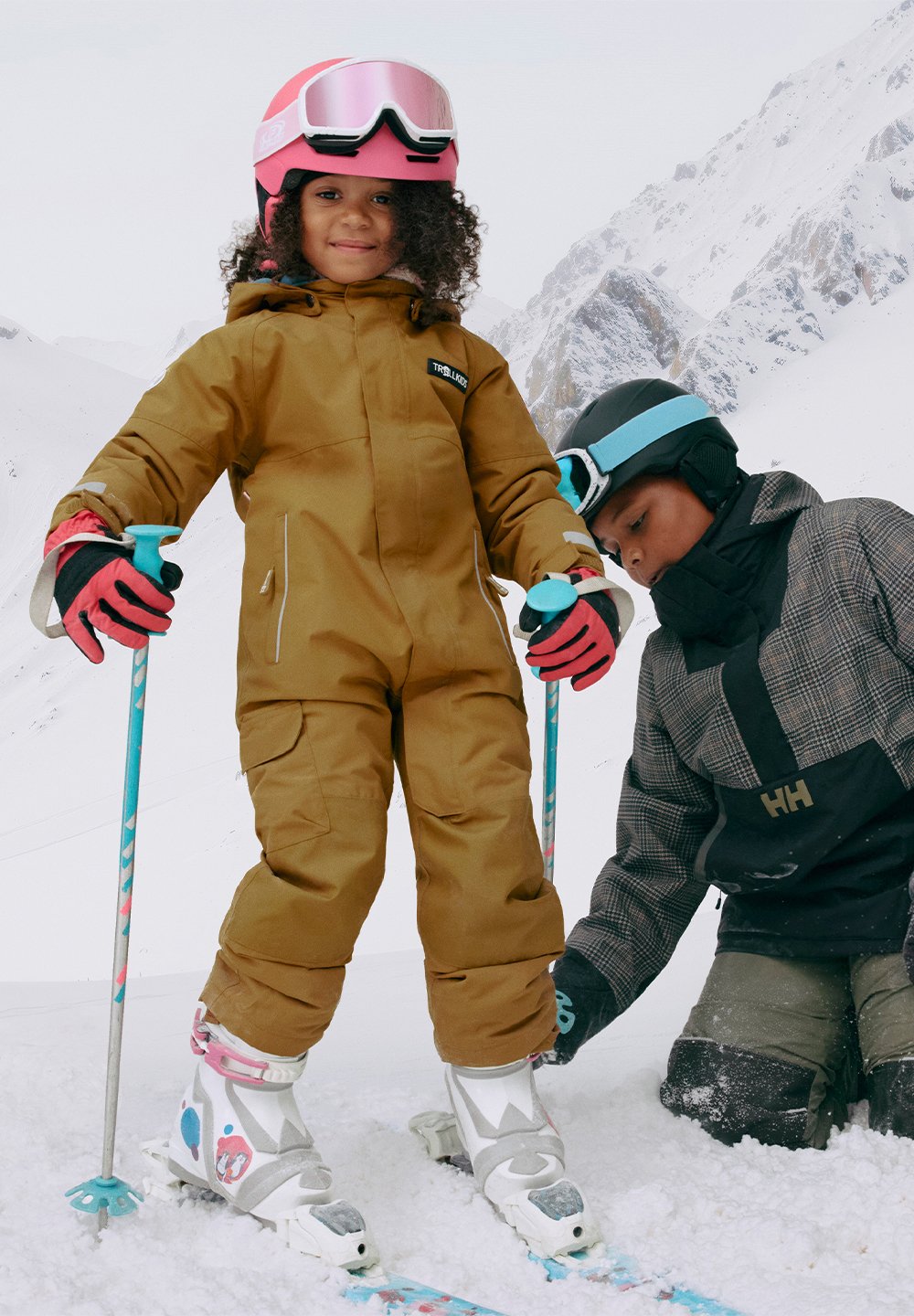 Barn i brun skidrakt med rosa hjelm og briller, står på ski og holder staver. Et annet barn justerer utstyr i nærheten. Snødekt bakgrunn.
