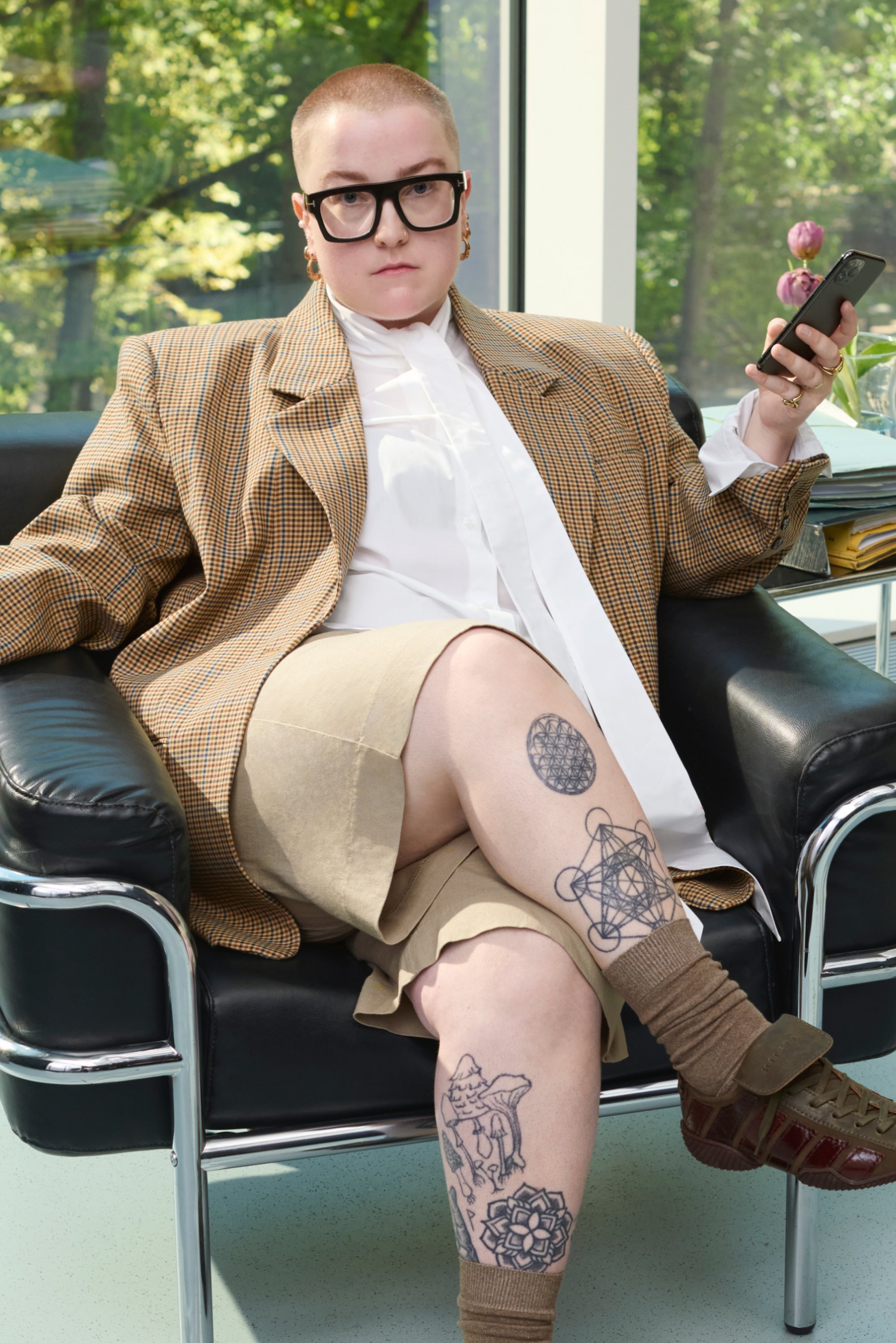 Persona che indossa un blazer oversize a quadri, una camicia bianca e pantaloni beige, seduta su una sedia in pelle nera con calze a fantasia e scarpe marroni.