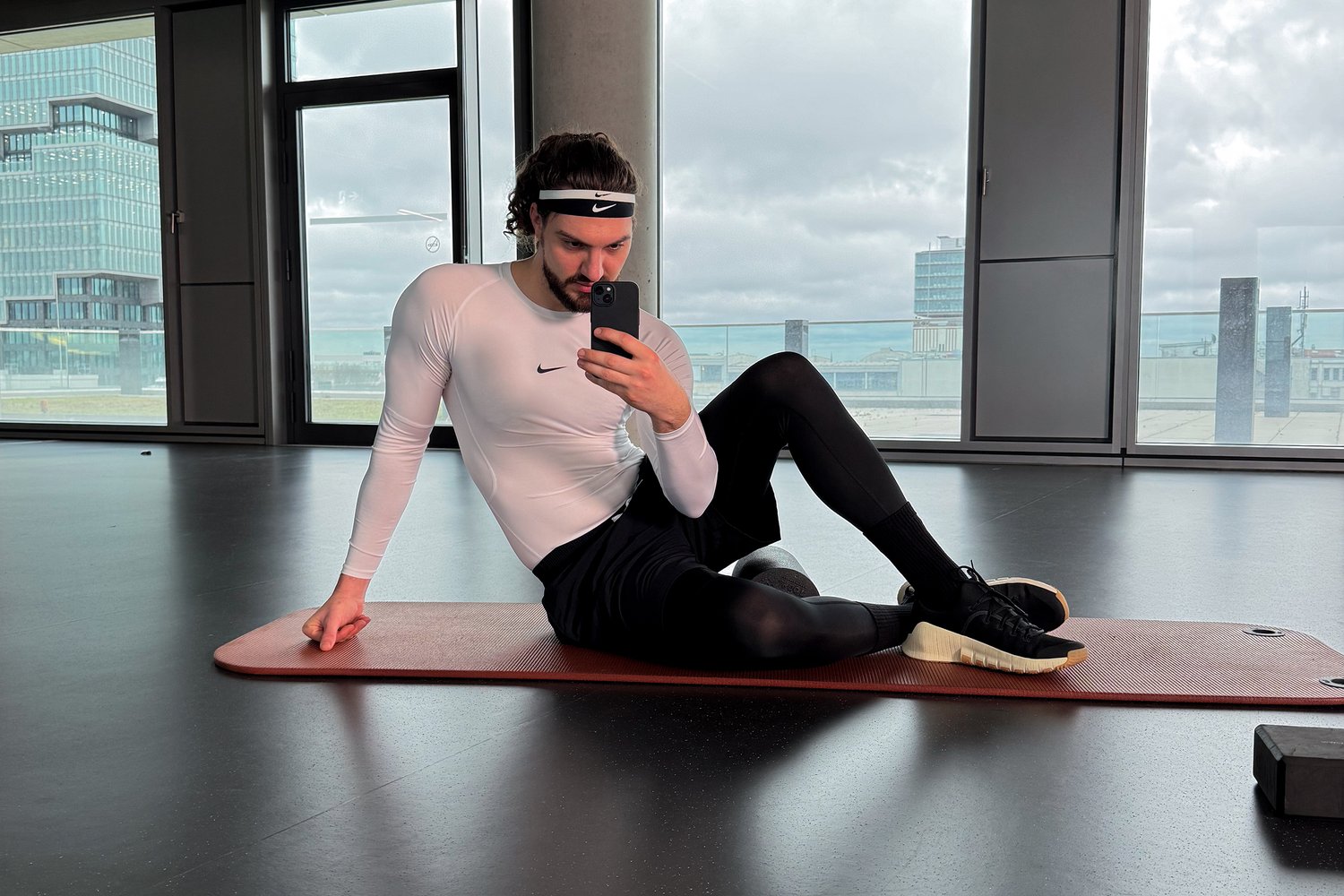 Un atleta masculino con una camiseta blanca de manga larga y mallas negras descansa sobre una esterilla roja en un gimnasio, tomándose un selfie con un teléfono inteligente. Grandes ventanas muestran una vista de la ciudad.