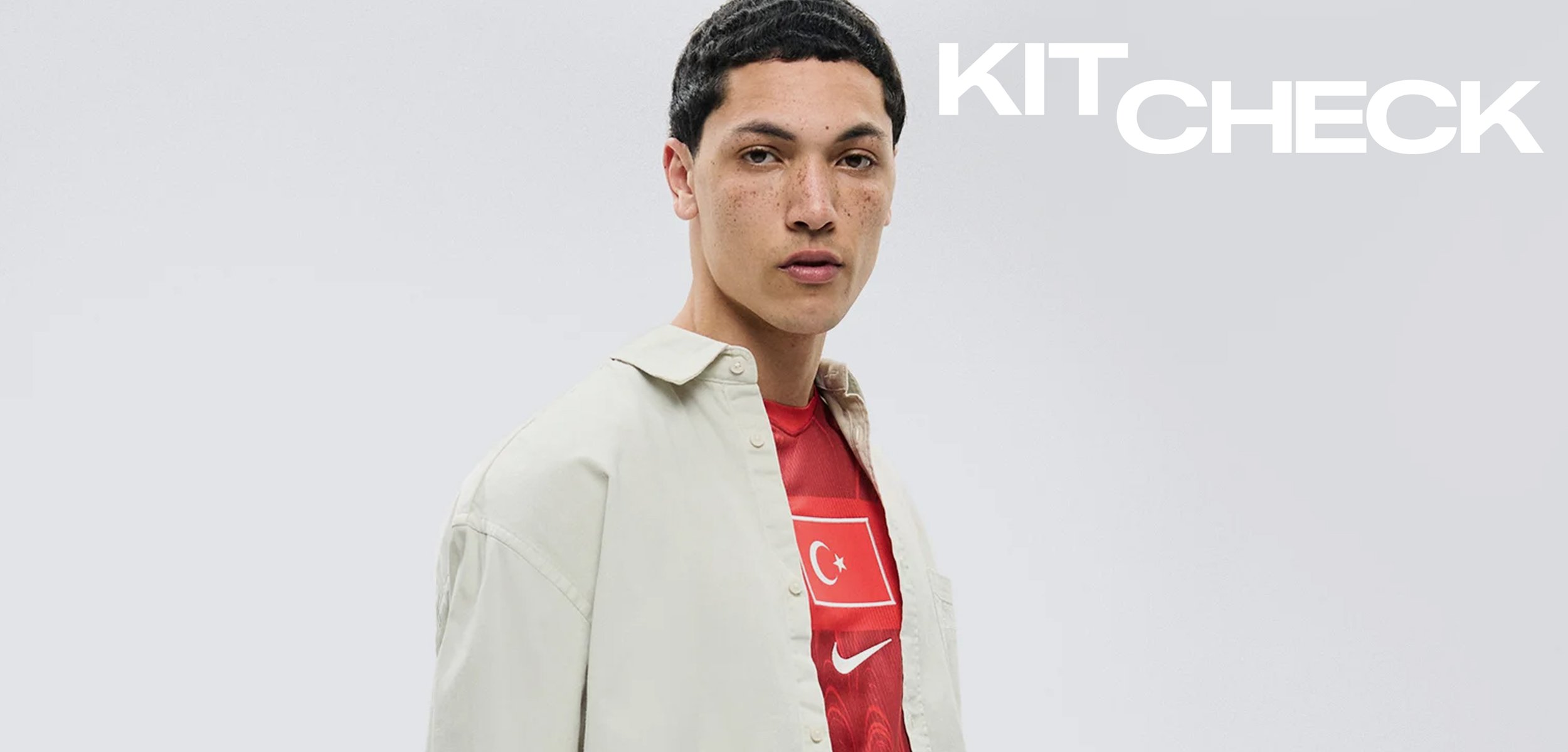 Mann mit kurzen schwarzen Haaren trägt ein rotes türkisches Fussballtrikot unter einem offenen beigefarbenen Hemd und steht vor einem schlichten hellen Hintergrund mit dem Text "KIT CHECK".