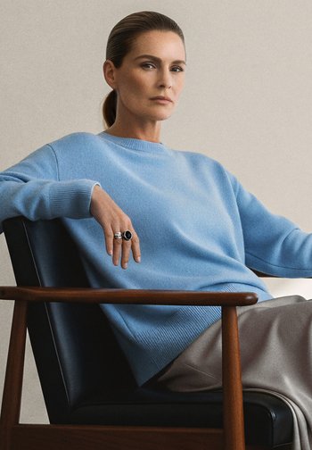 Lyseblå strikkegenser med ribbede mansjetter, båret over beige bukser. Modellen sitter i en trestol, iført sølvringer på hånden.