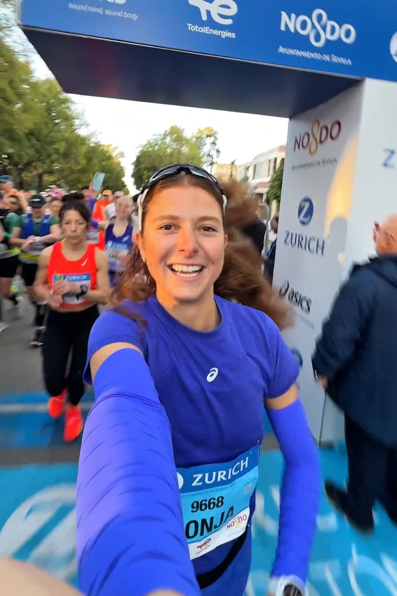 Lachende vrouwelijke hardloper in blauwe outfit met startnummer 9668 maakt een selfie bij de startlijn van een marathon, met andere hardlopers achter haar.