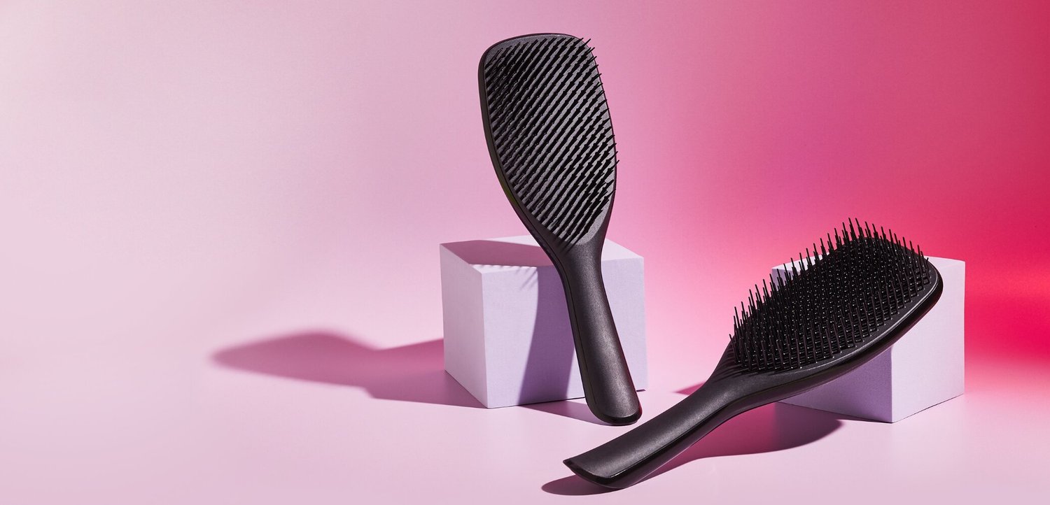 Deux brosses à cheveux noires avec des poils flexibles posées sur et près de cubes blancs sur un fond dégradé rose.