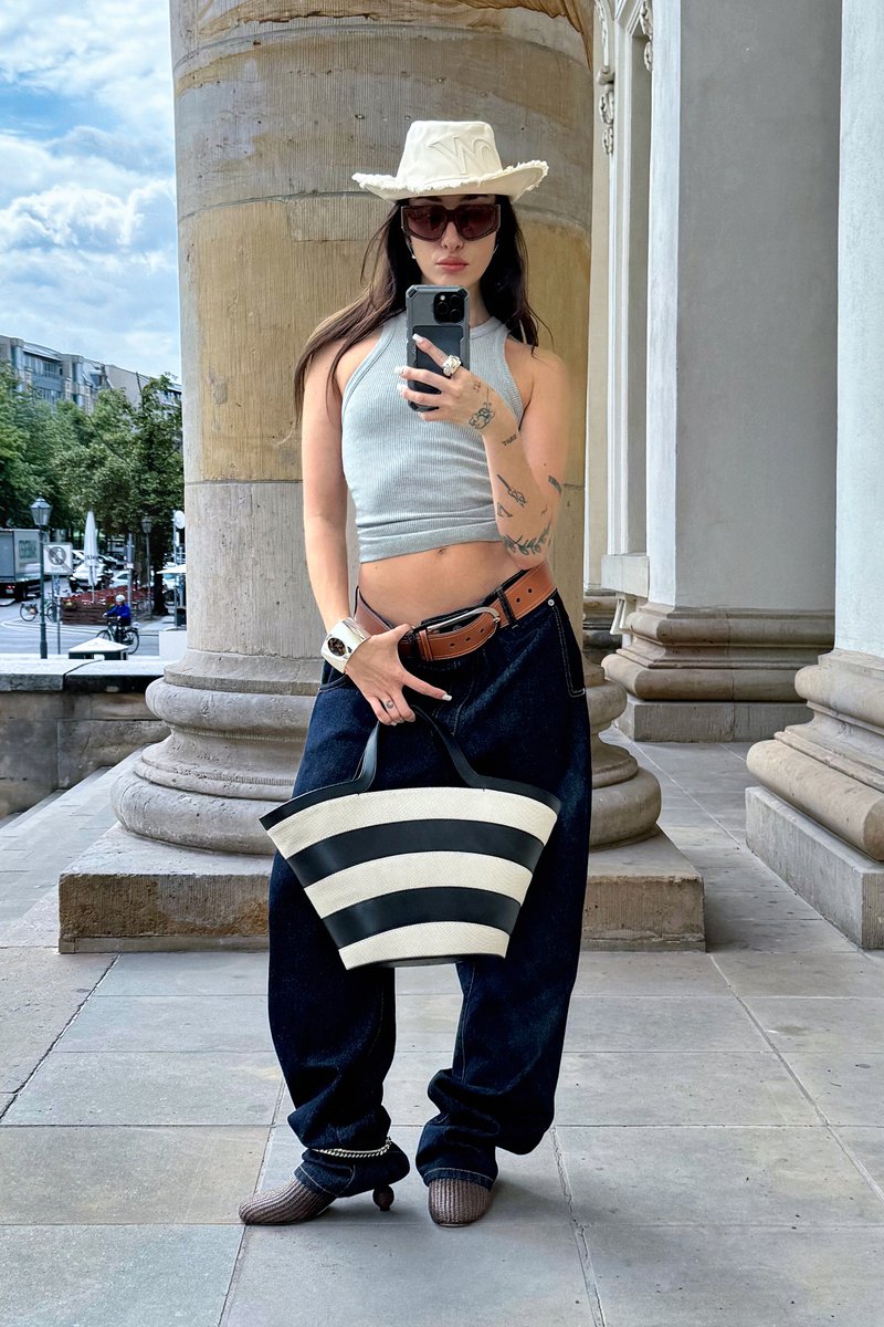 Borsa a strisce nera e bianca; top grigio corto; jeans larghi scuri; cappello bianco a tesa larga; occhiali da sole; in piedi su pavimento in pietra vicino a colonne.