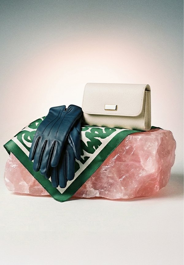 Guantes de cuero negros y una cartera beige descansan sobre un paño verde con estampado, encima de una gran roca de cristal rosa.
