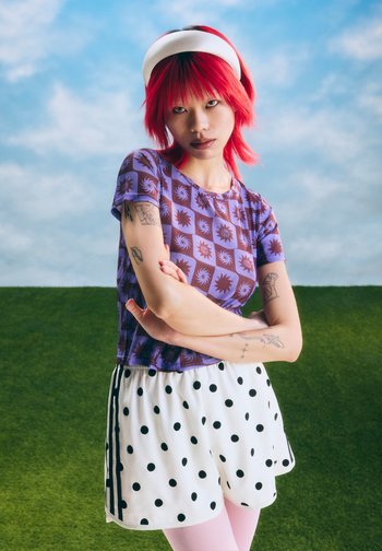 Jeune personne aux cheveux rouges vifs portant une chemise violette à motifs, un short blanc à pois et un bandeau blanc, debout devant une colline herbeuse.