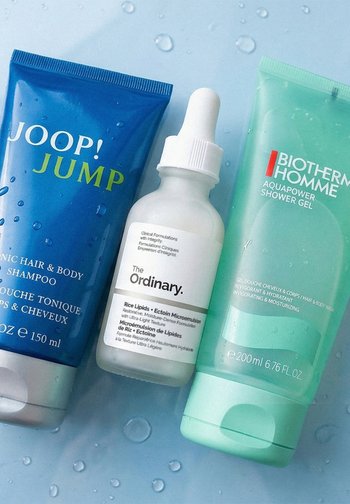 Tre hudplejeprodukter på en våd blå overflade: Joop! Jump shampoo, The Ordinary serumdråbe og Biotherm Homme shower gel.