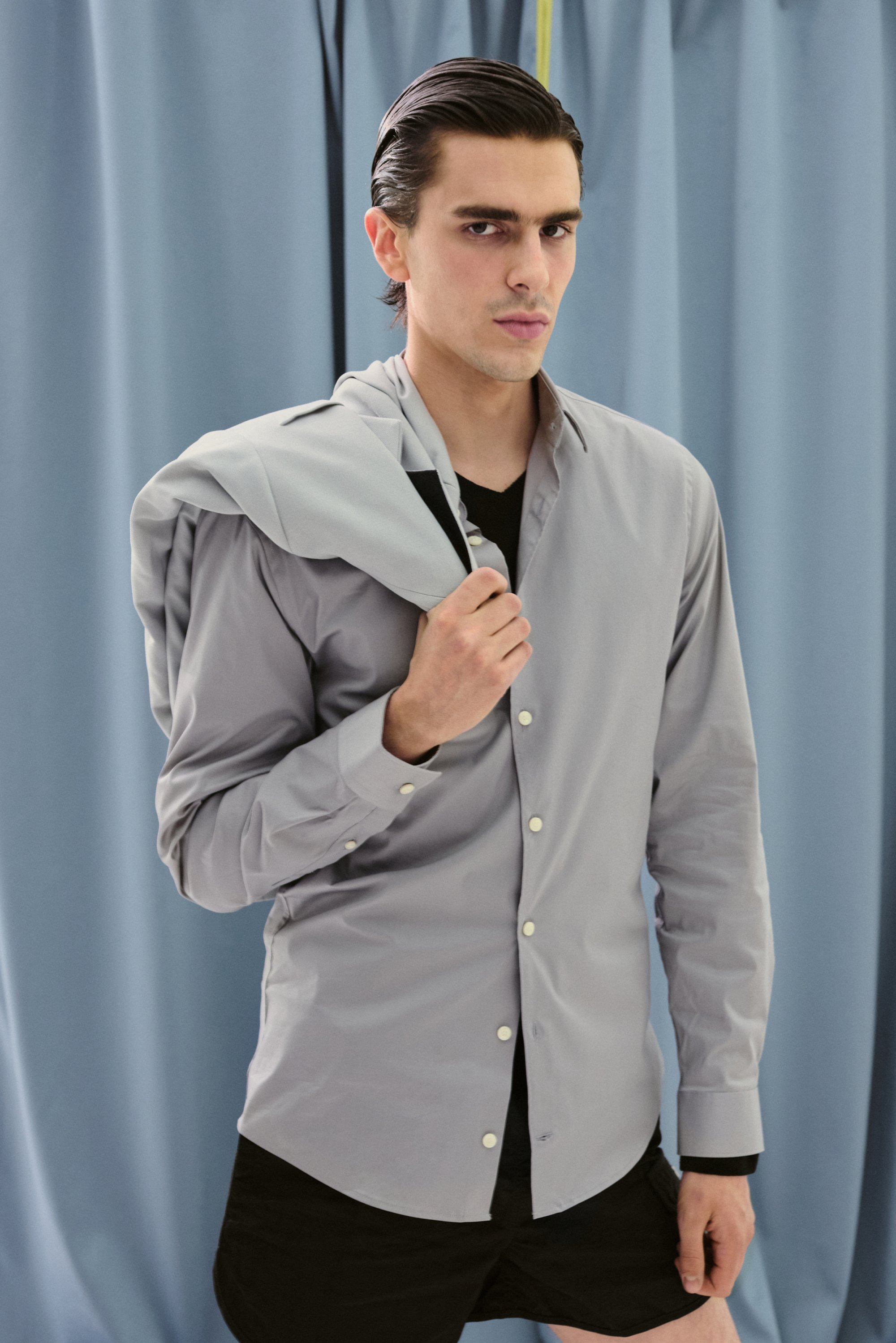 Un uomo con i capelli slicked-back sta in piedi con sicurezza di fronte a delle tende blu, indossando una camicia button-up grigia chiara con un hoodie drappato sulla spalla.