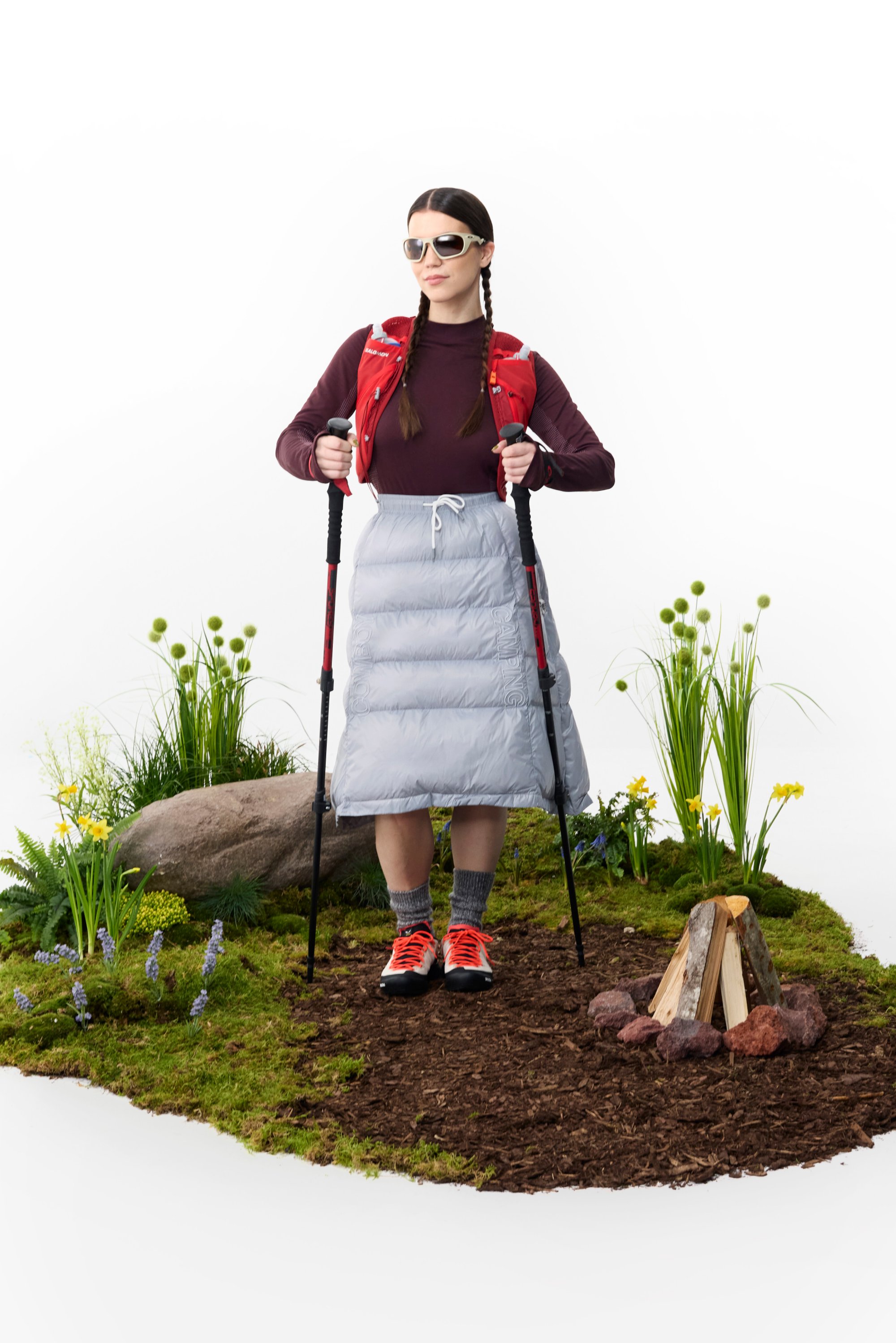 Una donna è in piedi su un prato, indossa un gilet rosso e una gonna imbottita grigia, mentre tiene dei bastoni da trekking accanto a un falò.