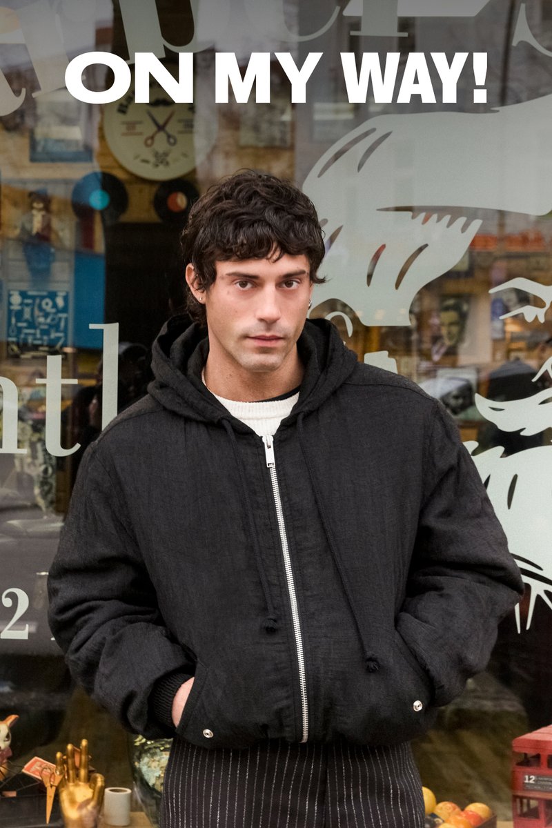 Un joven con una chaqueta negra está de pie fuera de una tienda de artículos vintage. Las palabras "¡EN CAMINO!" se muestran de manera prominente arriba.