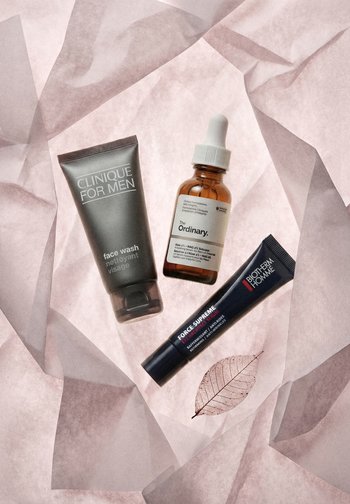Limpiador facial Clinique for Men, serum The Ordinary con cuentagotas y serum para ojos Biotherm Homme Force Supreme sobre papel arrugado de color rosa pálido con una hoja delicada.