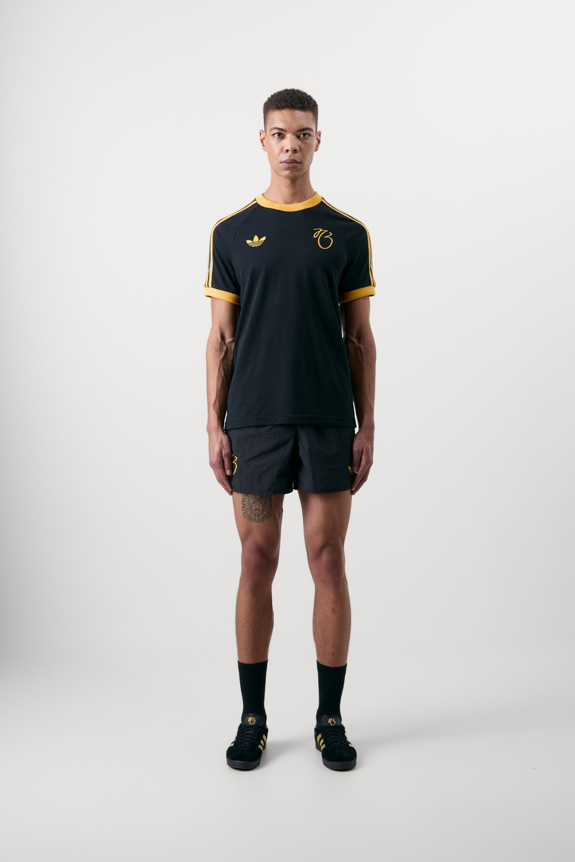 Una persona se encuentra de pie con confianza en un conjunto deportivo negro con acentos amarillos, que incluye un logo, pantalones cortos y zapatillas, sobre un fondo liso.