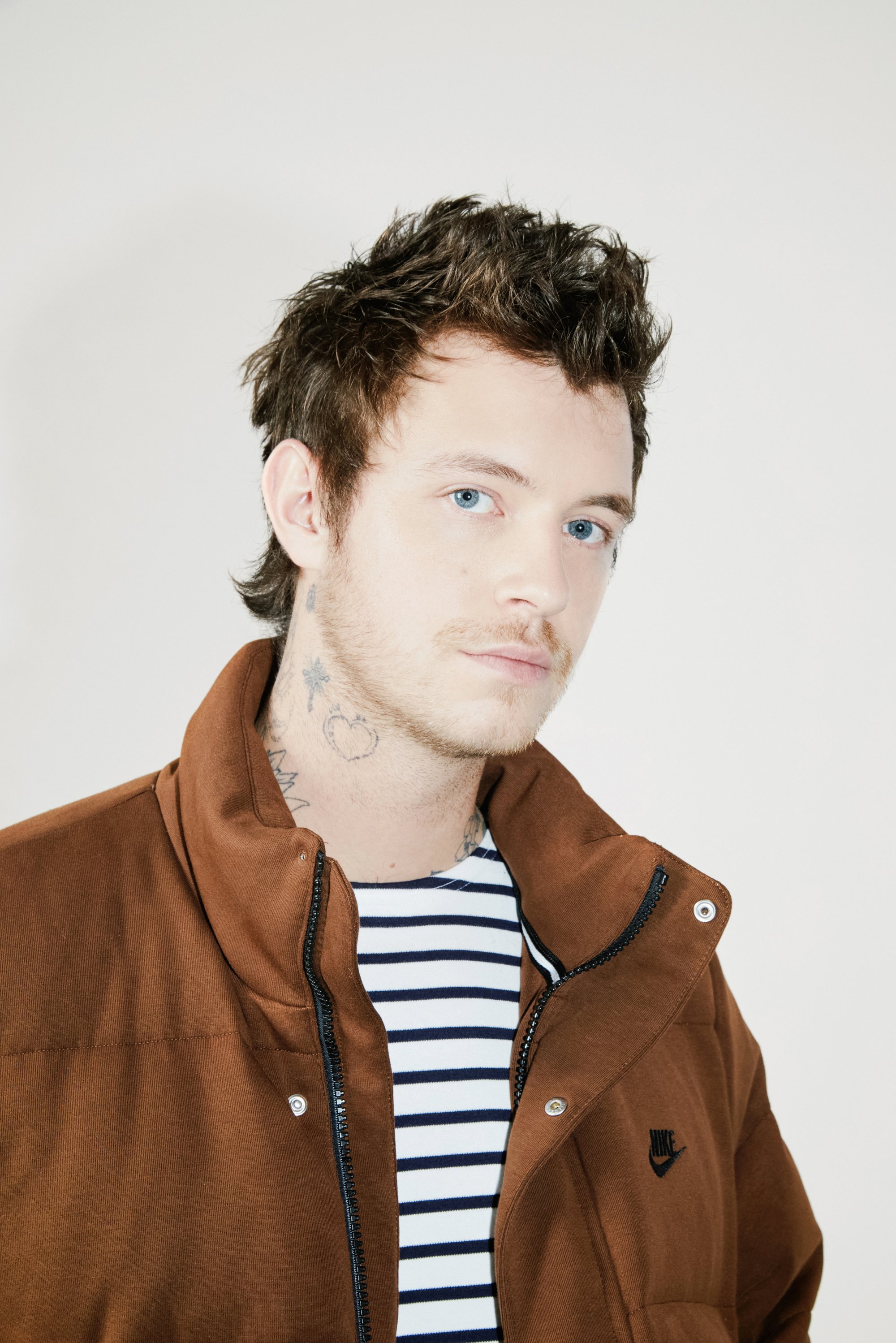 Un hombre con ojos azul claro y cabello corto y despeinado lleva una chaqueta marrón sobre una camisa a rayas, mostrando tatuajes en su cuello.