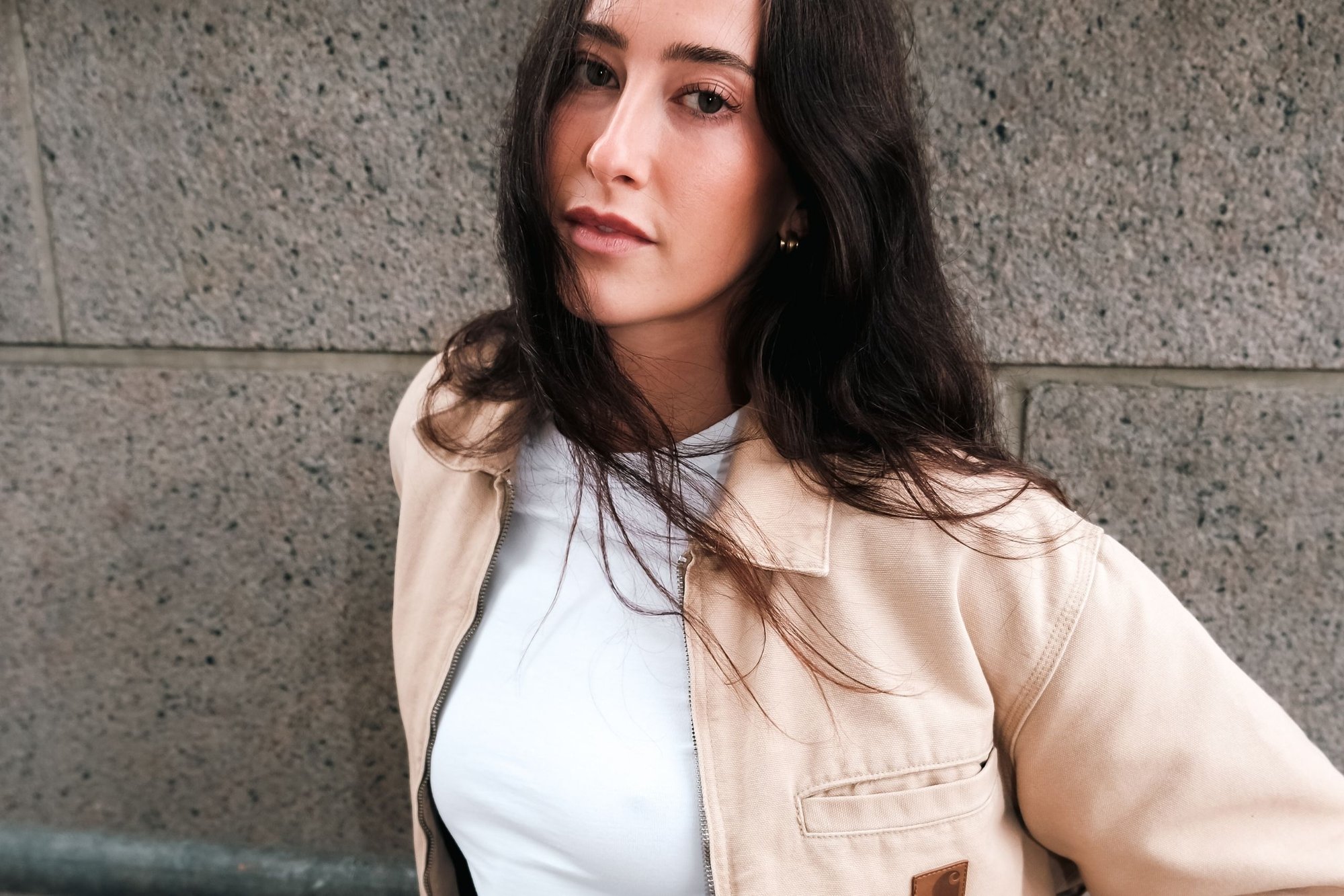 Beige, zippered jacka med en ljusfärgad T-shirt under, placerad framför en texturerad grå vägg. Jackan har en ficka och en logotyp. 