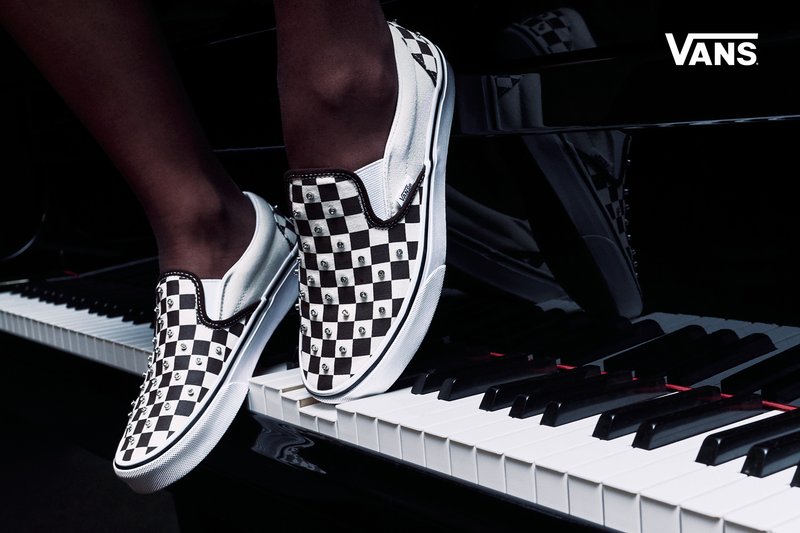 Person wearing Vans black and white checkerboard slip-on shoes standing on piano keys, Vans logo in top right corner.