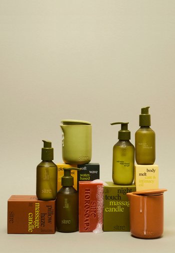 Zielone i pomarańczowe świece do masażu, butelki z olejkiem do masażu oraz czerwony kostka mydła z pianką ułożone na dopasowanych pudełkach na beżowym tle.