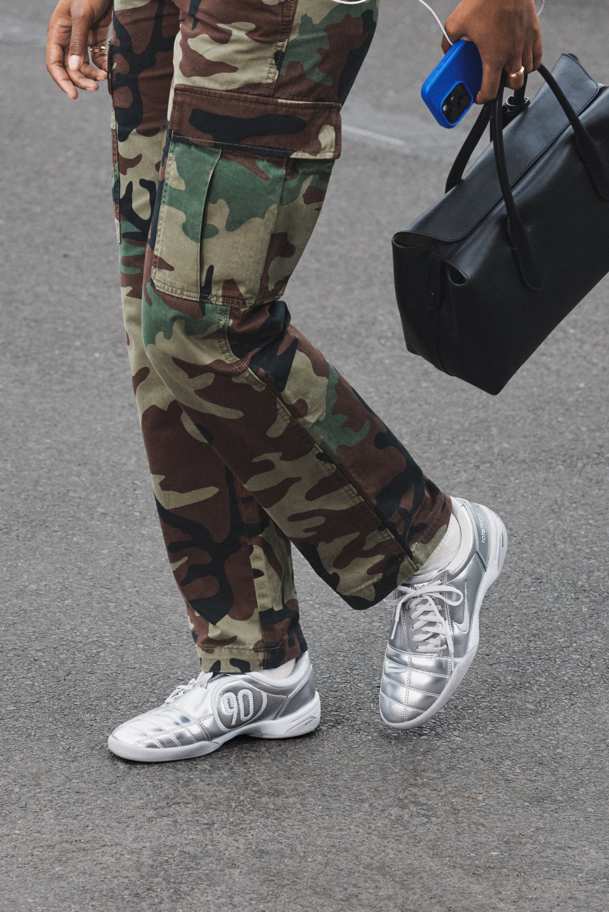 Zapatillas deportivas plateadas con una parte superior sintética texturizada, combinadas con pantalones cargo de camuflaje. Una bolsa negra y un teléfono azul visibles en la mano.