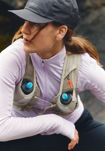 Kvinne med svart caps, lys lilla glidelåstop og drikkeryggsekk med vannflasker, ser mot venstre utendørs.