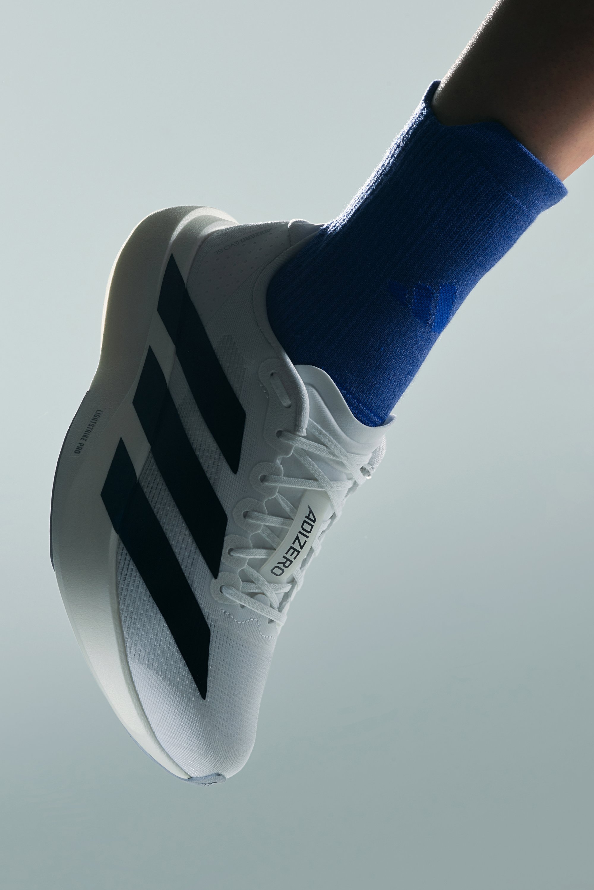 Zapatilla de running blanca con rayas negras y etiqueta "ADIZERO," llevada con un calcetín azul, contra un fondo gris suavemente iluminado.