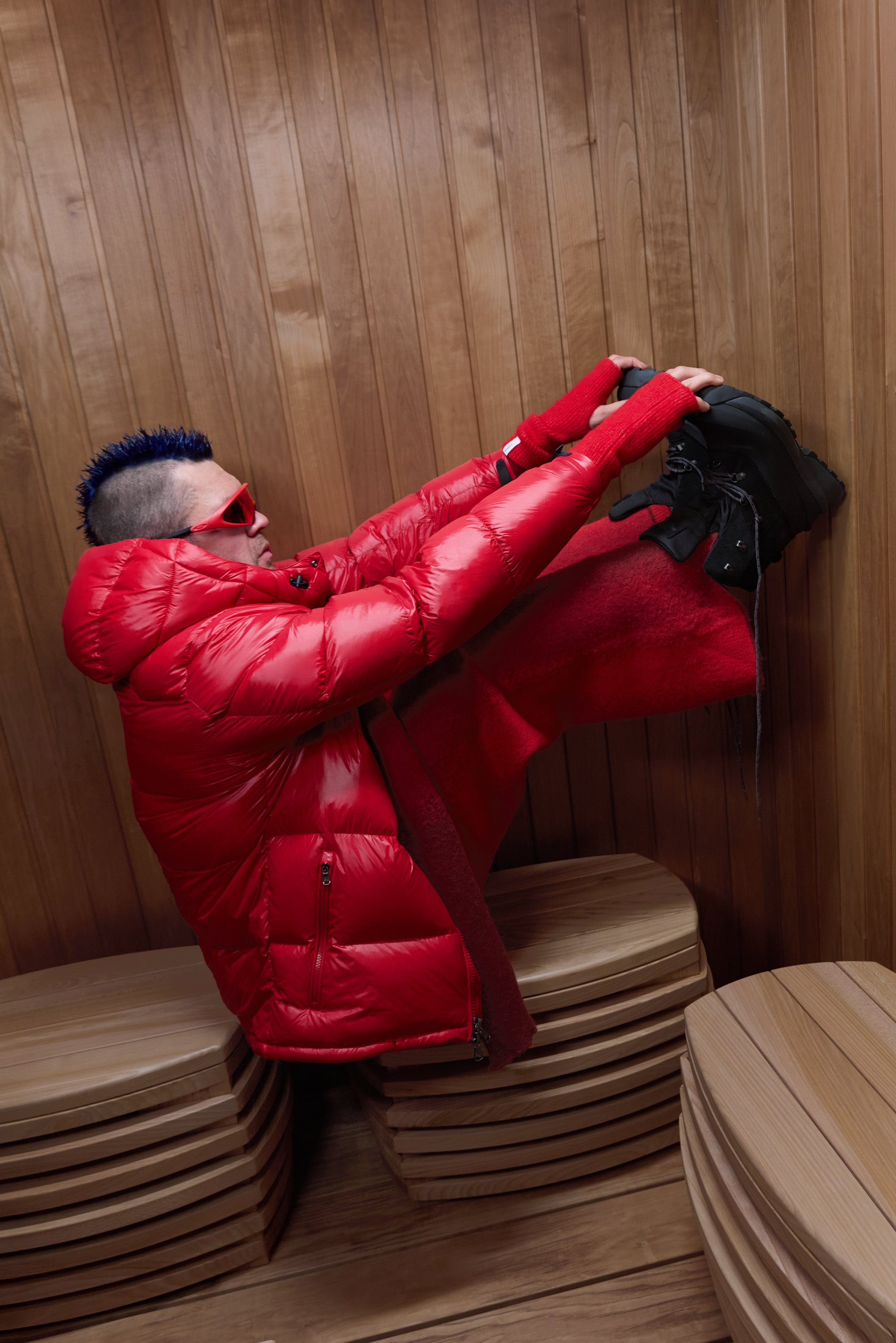 Punainen pöhö takki kiiltävällä tekstuurilla, mustat jalkineet ja punaiset sormettomat hansikkaat, asetettuna puiselle saunan sisustalle.