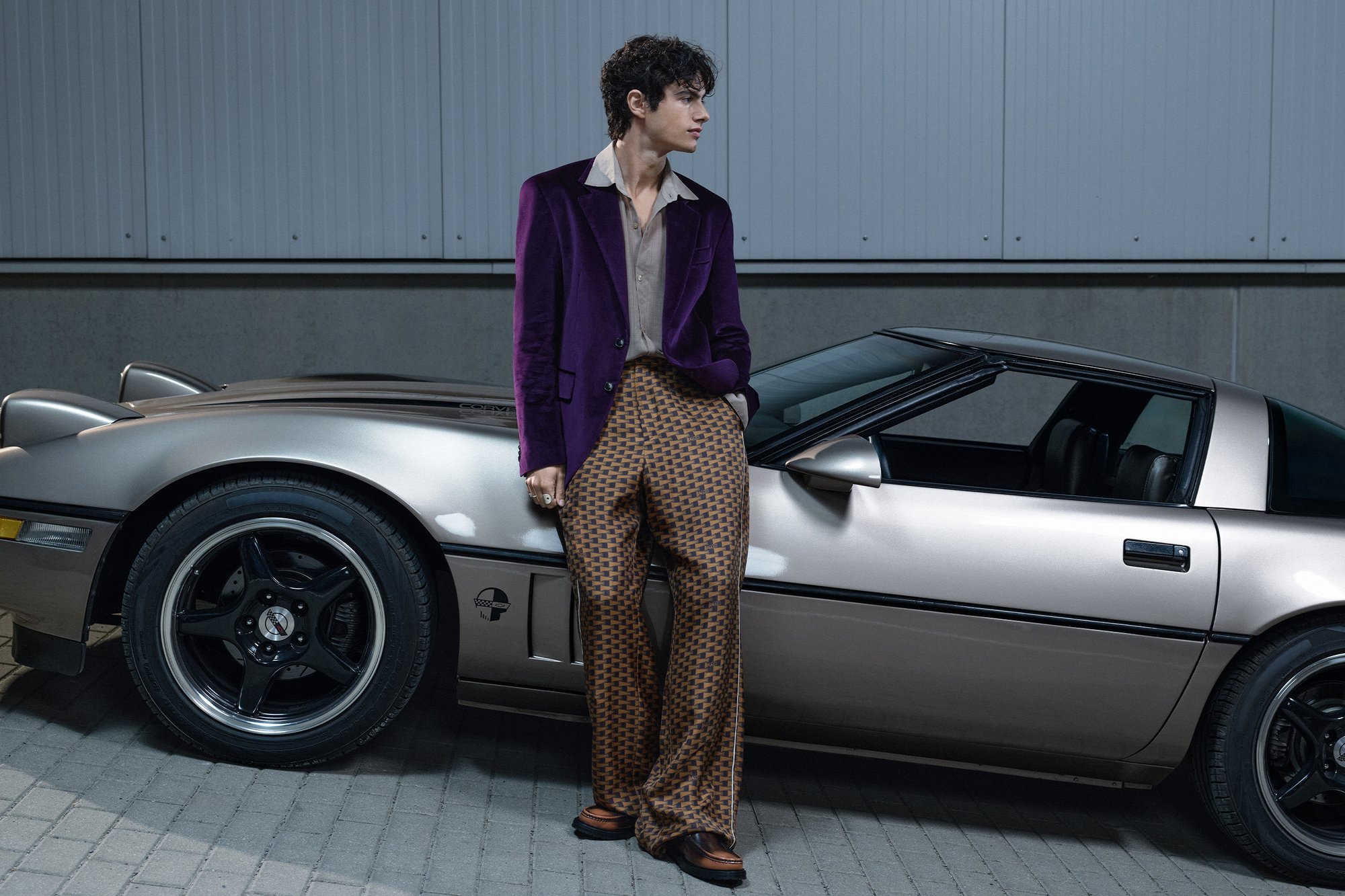 Una persona joven se encuentra de manera informal junto a un coche deportivo plateado, vistiendo una chaqueta morada y pantalones con patrón, con un fondo minimalista.