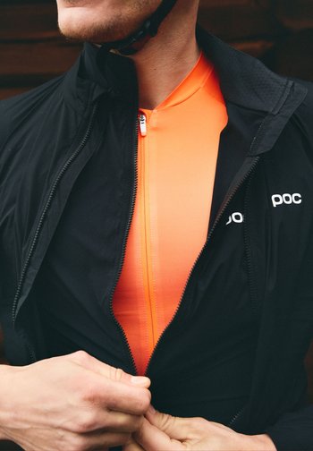 Person med en knall oransje sykkeltrøye under en delvis glidelåst svart POC-jakke, fester glidelåsen på jakken.