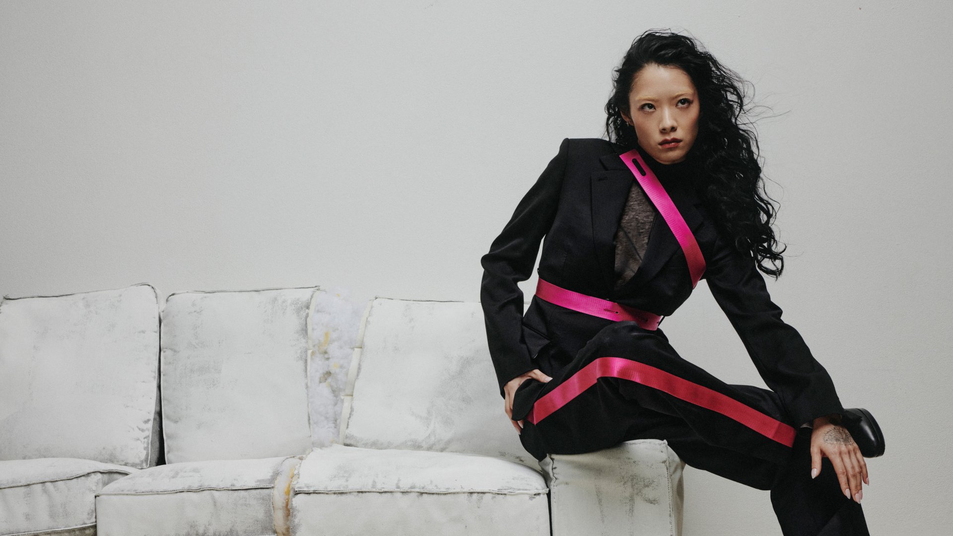 Una persona con cabello largo y negro está sentada en un sofá blanco desgastado, vistiendo un traje negro con acentos rosas y adoptando una pose fuerte frente a una pared lisa.