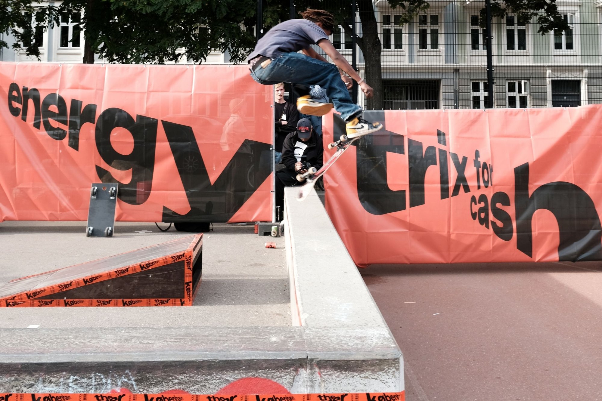 Skateboardramp på ett evenemang med en orange banderoll. En åkare i luften, iklädd en blå tröja och jeans, utför ett trick ovanför en betongbarriär.