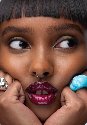 Gros plan d'une personne à la peau foncée, lèvres rouge foncé brillantes, piercing au septum, fard à paupières vert, et mains pressant les joues portant des bagues en argent et turquoise.