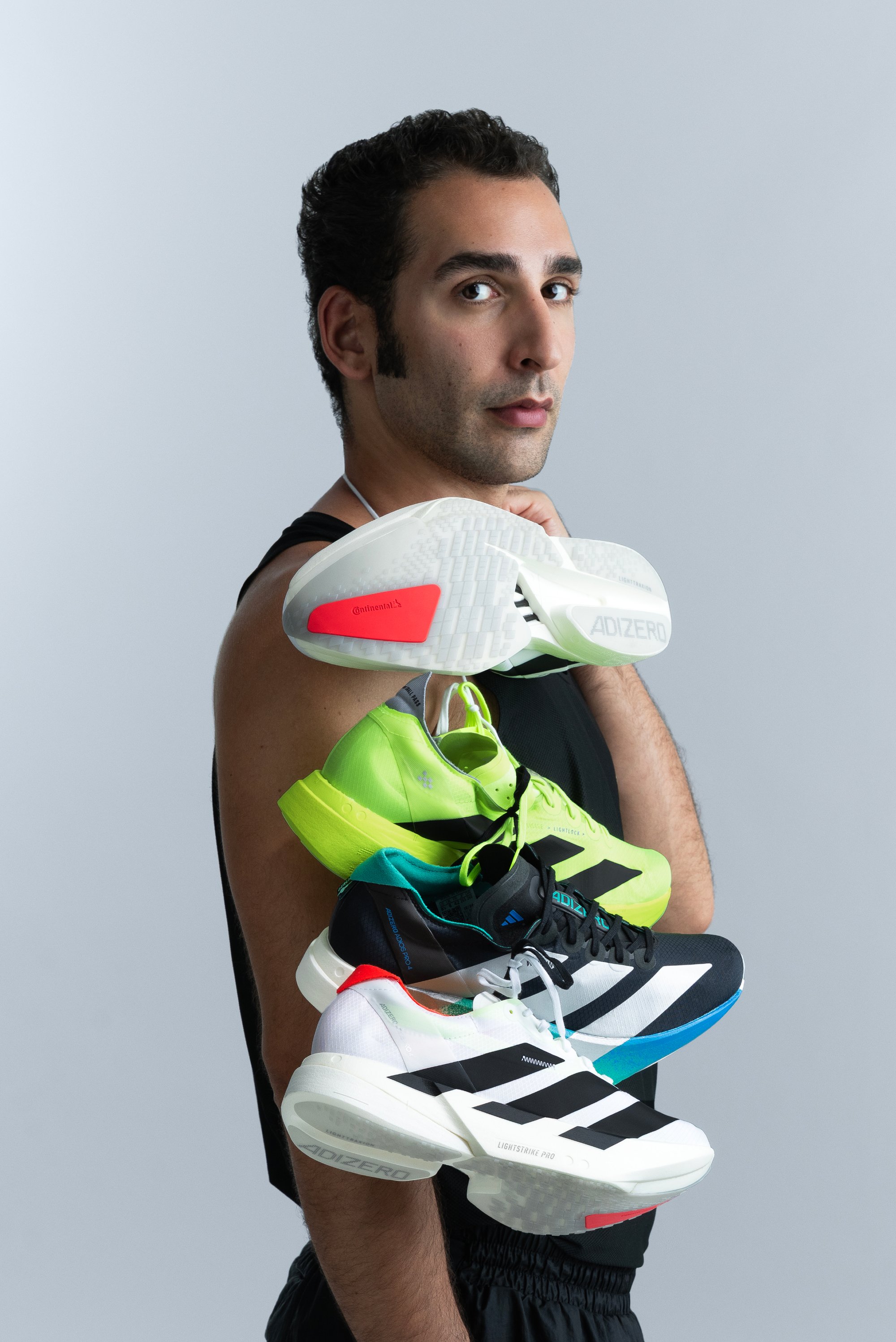 Homme en débardeur noir tenant trois chaussures de course Adidas de couleurs distinctes sur son épaule, devant un fond gris clair.