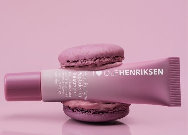 Różowa tubka kosmetyków Ole Henriksen ustawiona między dwoma fioletowymi makaronikami na jednolitym różowym tle.