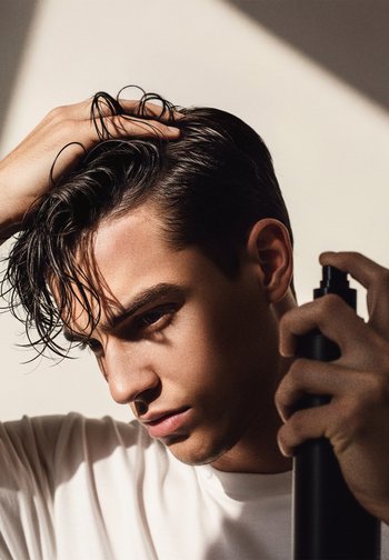 Bouteille vaporisateur noire tenue dans une main, avec des cheveux foncés brillants, coiffés et humides. Gros plan sur un jeune homme en chemise blanche, avec une légère ombre.