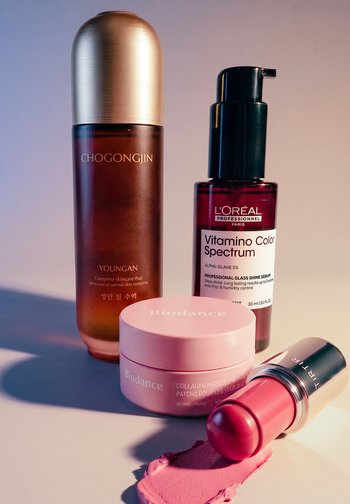 Cztery produkty do pielęgnacji skóry i makijażu: brązowa butelka z serum, czerwona butelka z pompką, pojemnik z różowymi plastrami kolagenowymi pod oczy oraz różowa szminka z rozmazaniem.