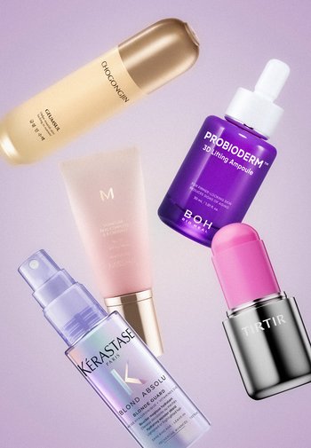 Cinq produits de soin et de beauté, dont des flacons, un tube et un spray, disposés en diagonale sur un fond violet clair. Aucune personne présente.
