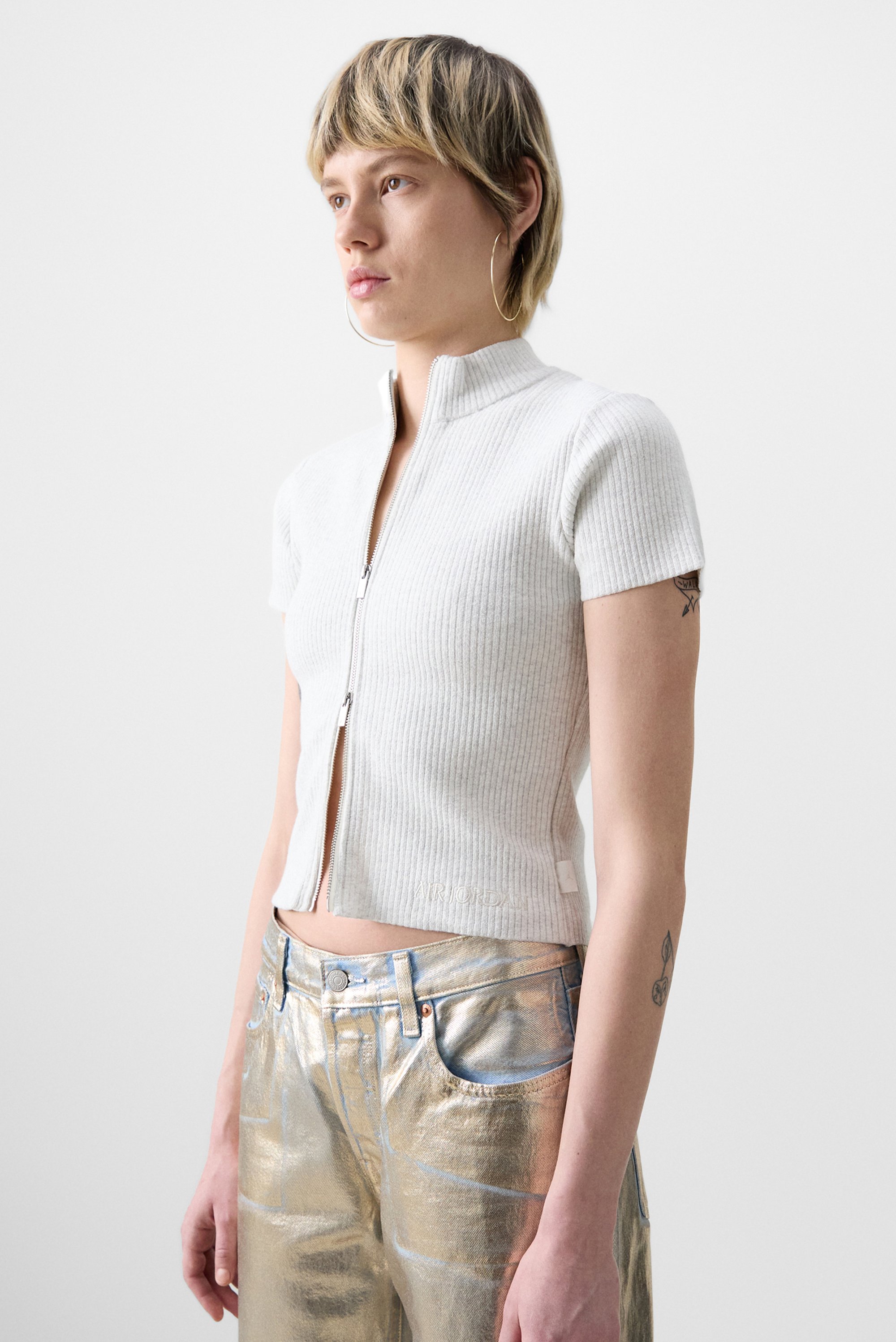 Persona con capelli corti e texturizzati che indossa un top bianco con zip e pantaloni metallici argentati lucenti, in piedi contro uno sfondo chiaro.