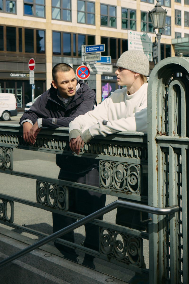 Due giovani che si appoggiano a una ringhiera decorativa in metallo lungo una strada di città, con segnali stradali e edifici moderni sullo sfondo.