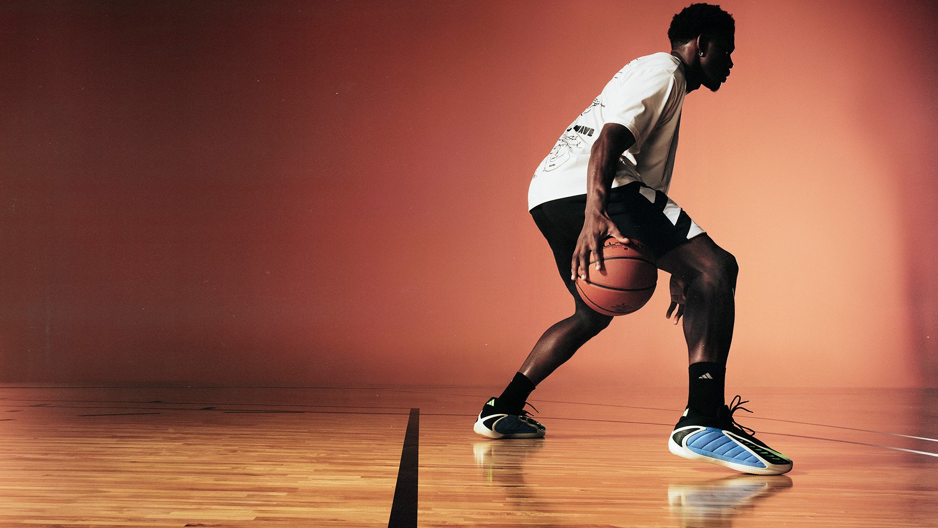 Giocatore di basket che dribbla una palla, indossando una maglietta grafica bianca, pantaloni corti neri con dettagli bianchi e scarpe blu e nere a fantasia.