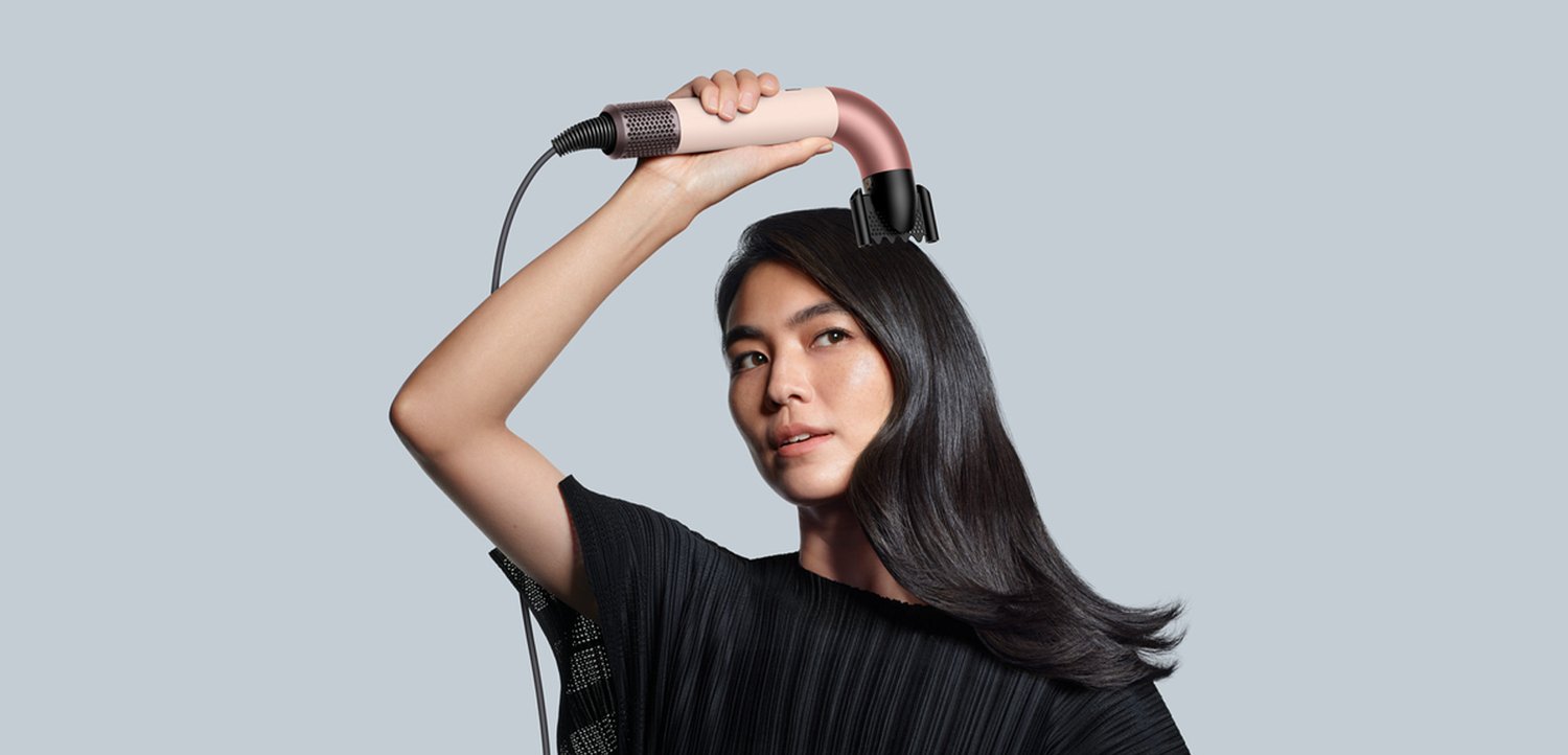 Een gebogen haarstylingtool met een roze en zwarte design, vastgehouden boven het hoofd met een lange kabel, tegen een lichtgrijze achtergrond.