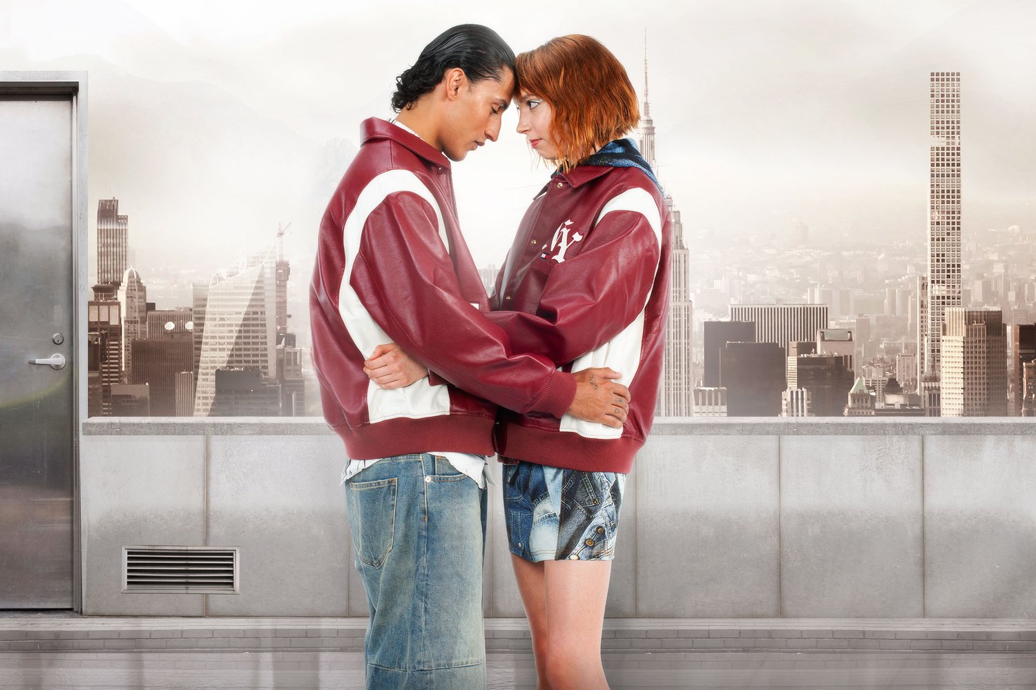 A man and woman in matching burgundy jackets embrace on a rooftop, with a city skyline and skyscrapers in the background.
