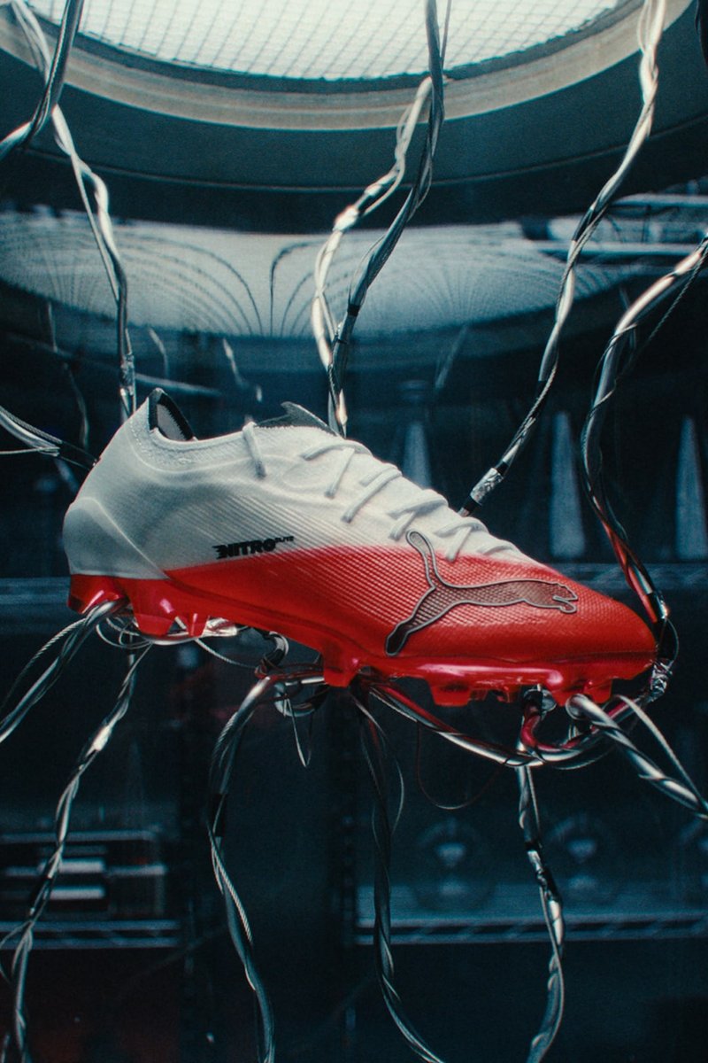 Weißer und roter Puma-Fußballschuh, verbunden mit mehreren schwarzen Kabeln in einer dunklen, industriellen Umgebung mit einem Gitterdecke darüber.