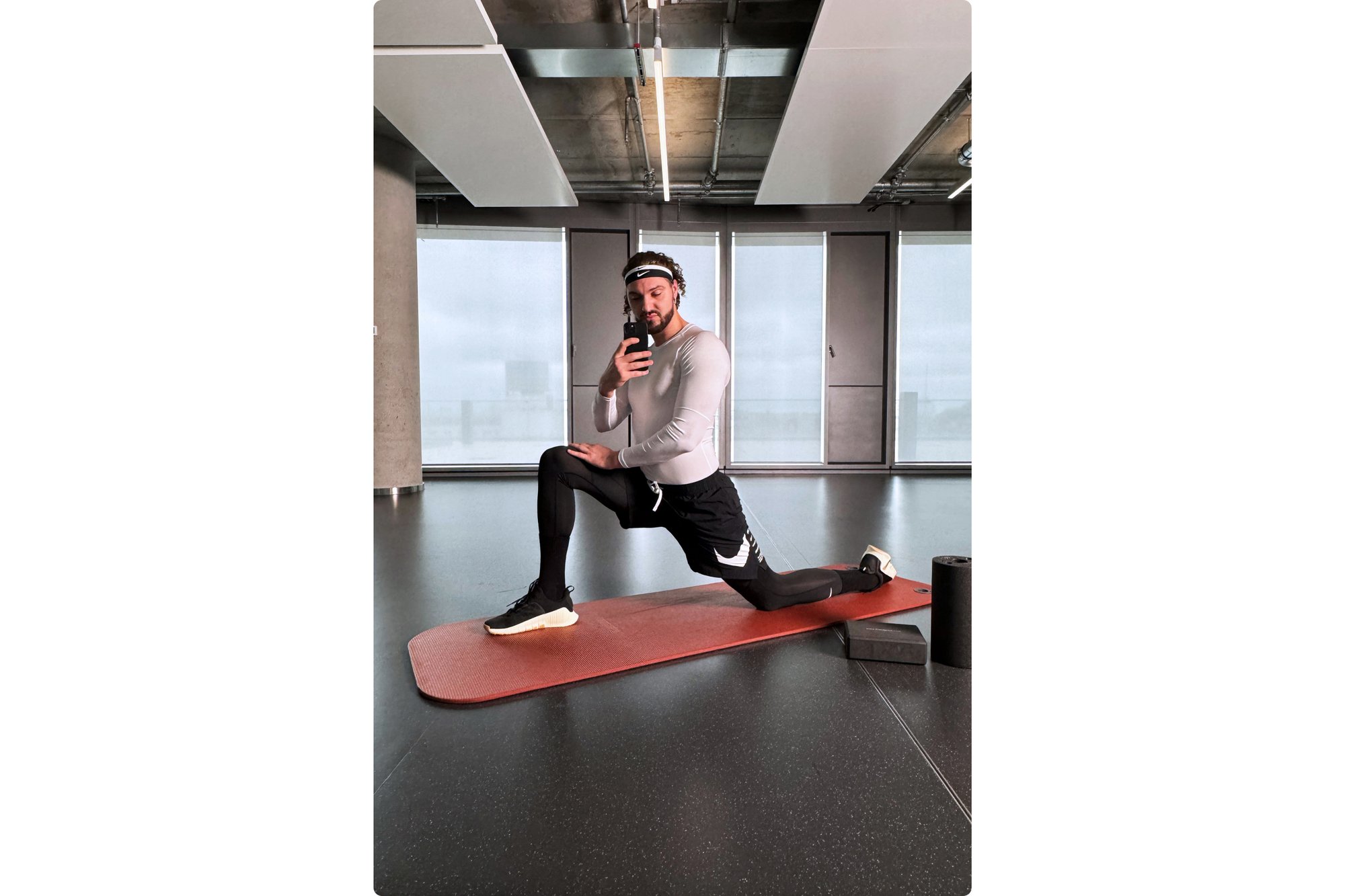 Una persona con ropa deportiva realiza una zancada sobre una esterilla de yoga en un estudio espacioso. Espejos reflejan grandes ventanas con una vista nublada del exterior.