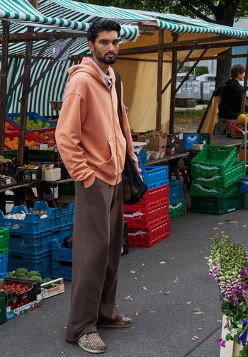 Model draagt een licht perzikkleurige hoodie, een paarse broek met wijde pijpen en schoenen met een luipaardprint, terwijl ze een zwarte mesh tas draagt op een markt.
