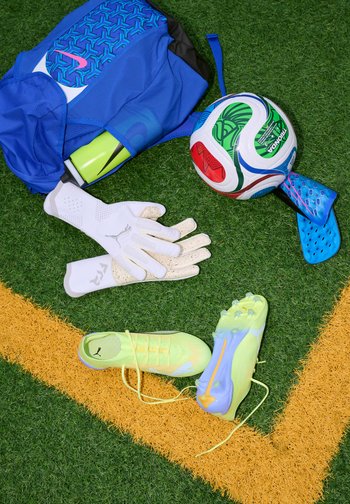 Zila sporta soma ar ūdens pudeli, baltas vārtsarga cimdi, futbola bumba, zili kāju aizsargi un dzelteni futbola apavi uz mākslīgā zālāja.