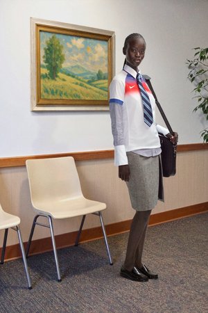 Camisa Nike blanca y azul con acentos rojos, mangas a rayas, falda lápiz gris, medias marrones, mocasines negros y un bolso de hombro.