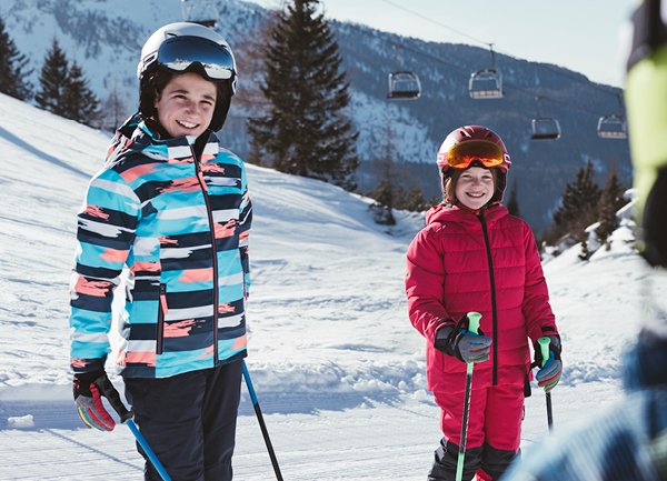 To barn iført fargerike skijakker står på snø, den ene i en blå og rosa stripete jakke, den andre i en rød, polstret jakke, med skistaver.