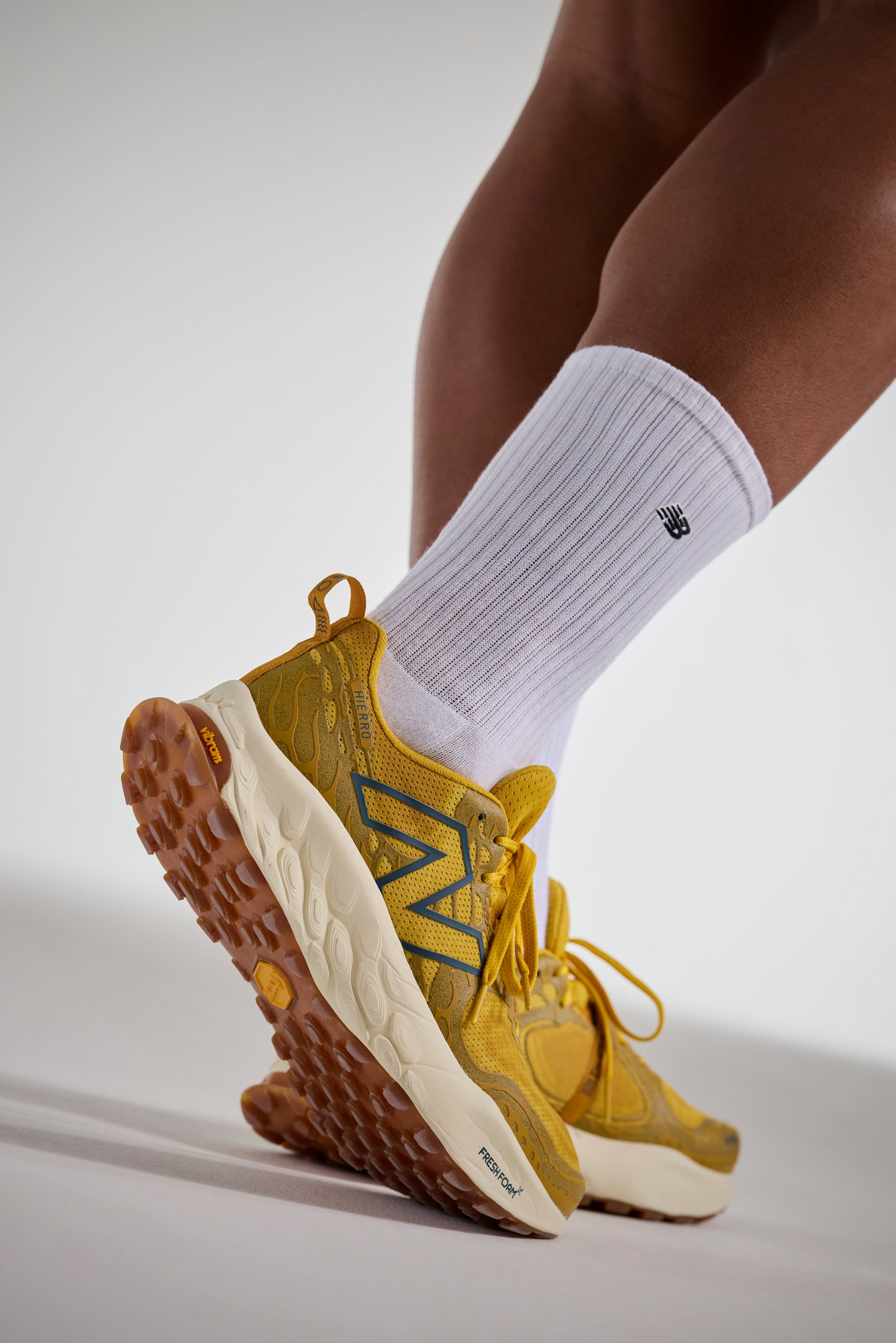 Primer plano de un pie que lleva una zapatilla amarilla de New Balance y un calcetín blanco acanalado, destacando los detalles del calzado sobre un fondo neutro.