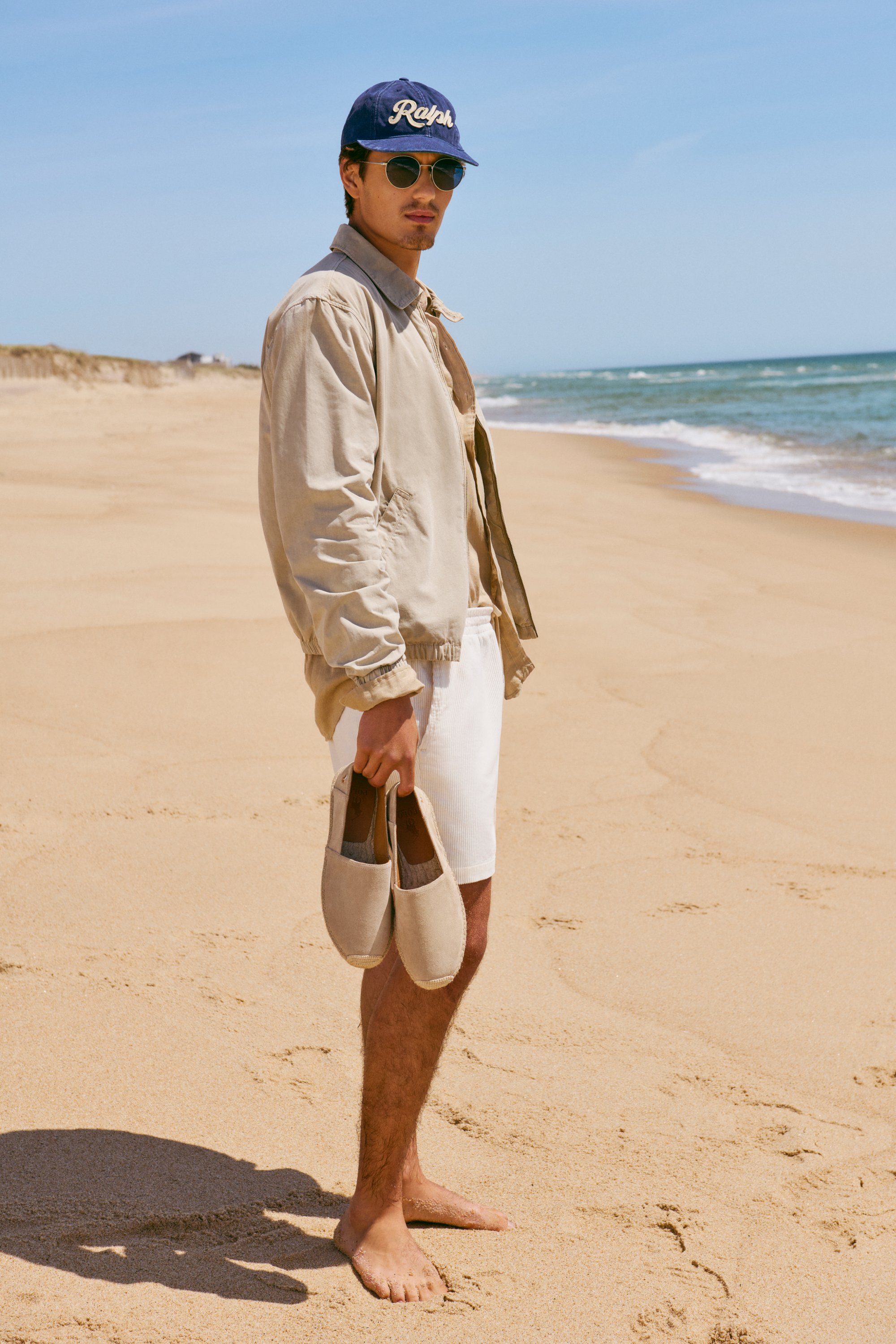 Un joven está descalzo en una playa de arena, sosteniendo zapatos beige. Lleva una chaqueta ligera, pantalones cortos blancos, gafas de sol y una gorra azul.