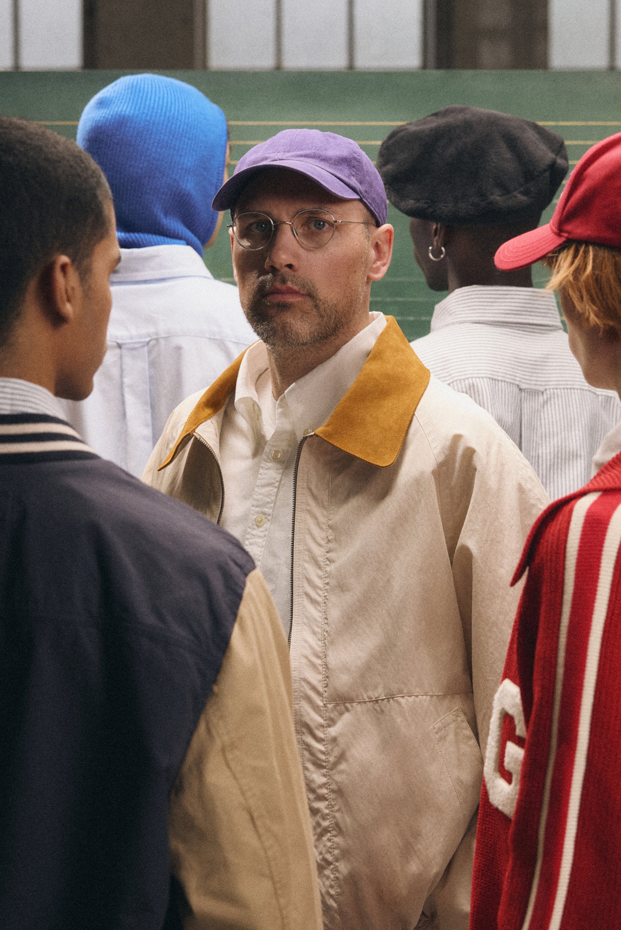 En man med glasögon och en lila keps står i fokus bland flera andra i avslappnad klädsel, mot en bakgrund av gröna linjer.