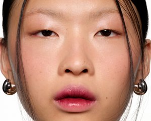 Primo piano di un volto con pelle liscia, lentiggini sottili, occhi scuri e un colore delle labbra sfumato. Indossa un orecchino a cerchio in argento.