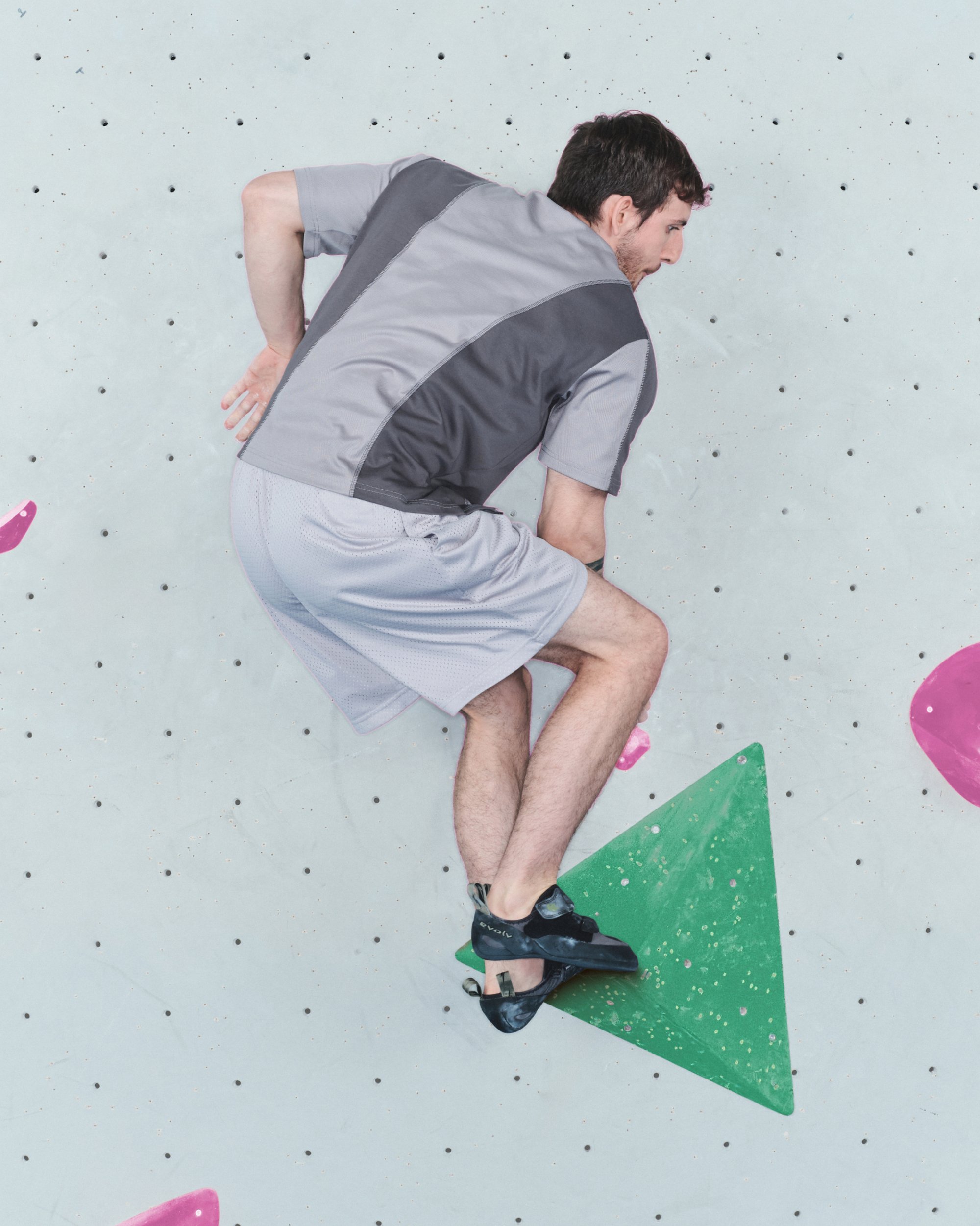 Escalador vistiendo una camiseta de manga corta gris y negra y pantalones cortos gris claro. Posicionado en un agarre triangular verde contra una pared texturizada.